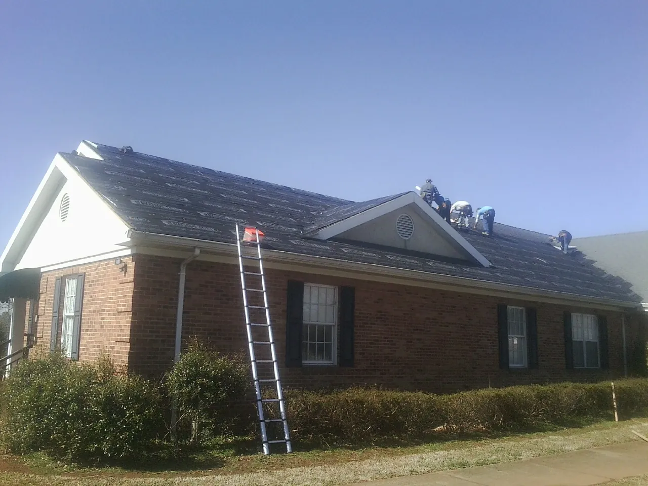 Crew tearing shingles off a brick ranch roof with a ladder set — Emergency Roof Repair work by our crew in Roca