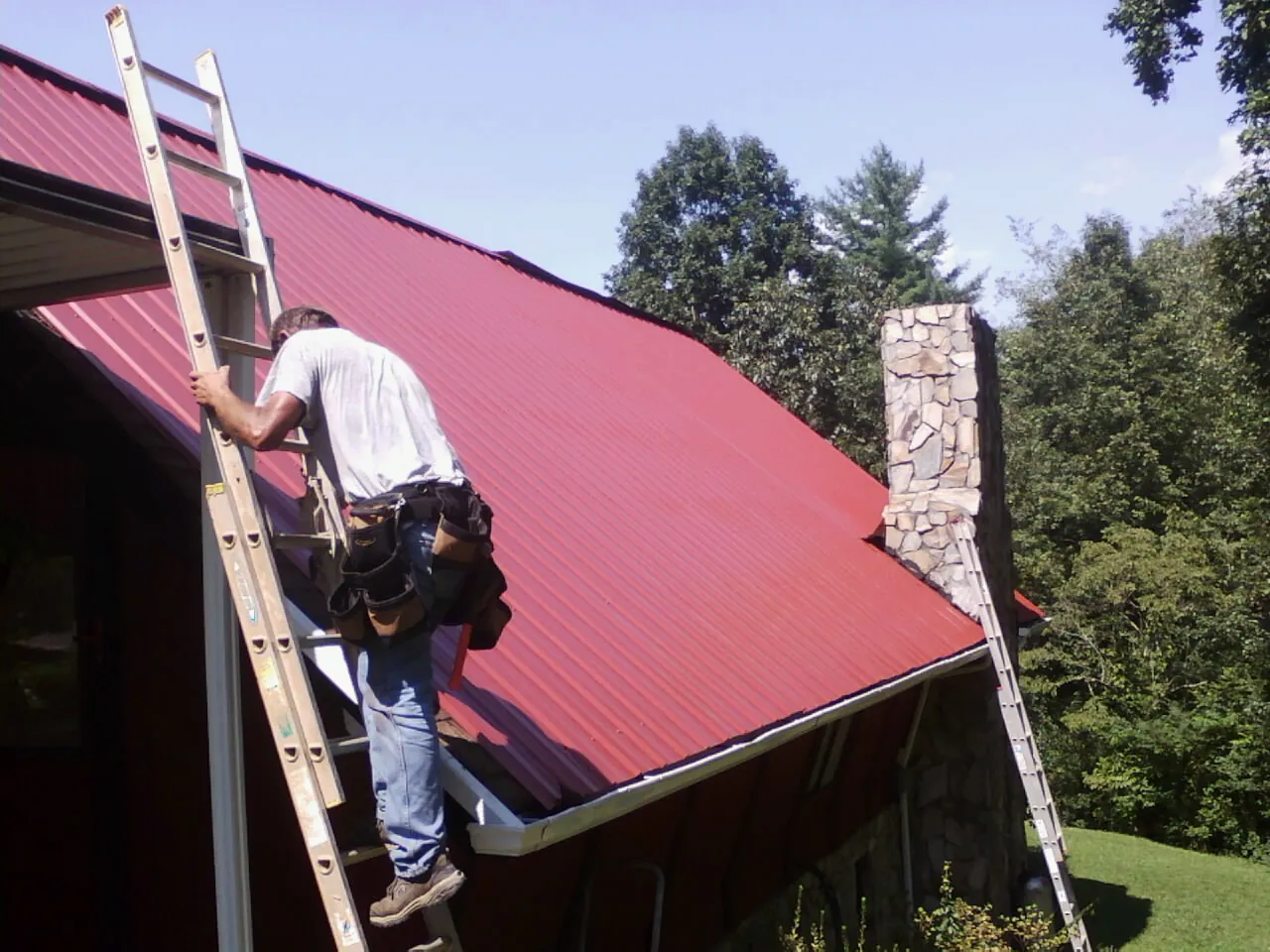 Roofer on a ladder beside a red metal roof and stone chimney — Soffit & Fascia Repair by Roofing Brothers in Roca